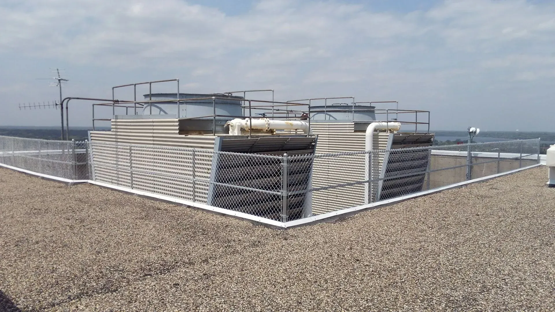 Rooftop cooling system with safety railings on a flat, gravel-covered roof against a cloudy sky.