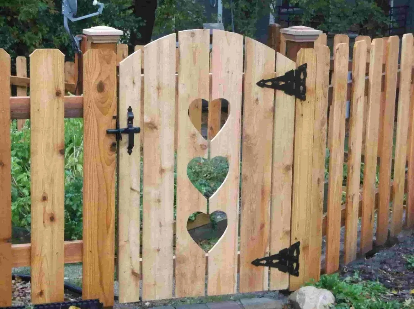 Wooden gate with three heart-shaped cutouts, part of a matching light brown picket fence. Black hinges and latch.