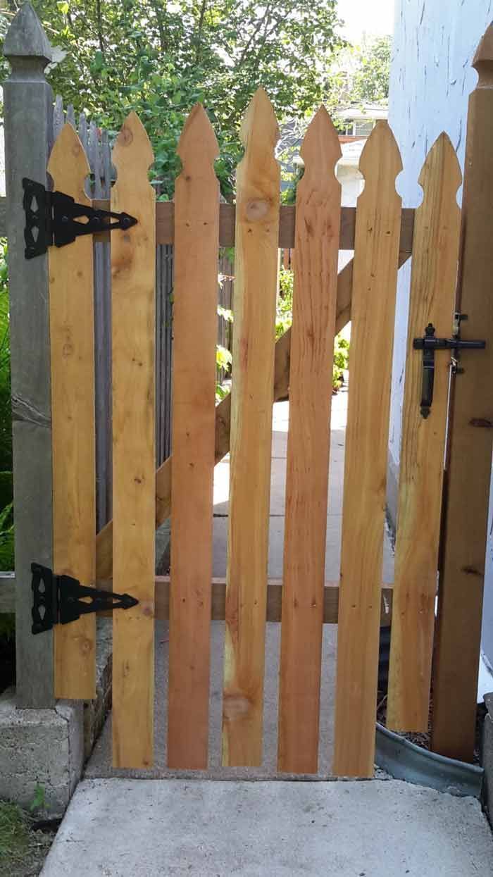 A wooden gate with black hinges is sitting on a sidewalk next to a fence.