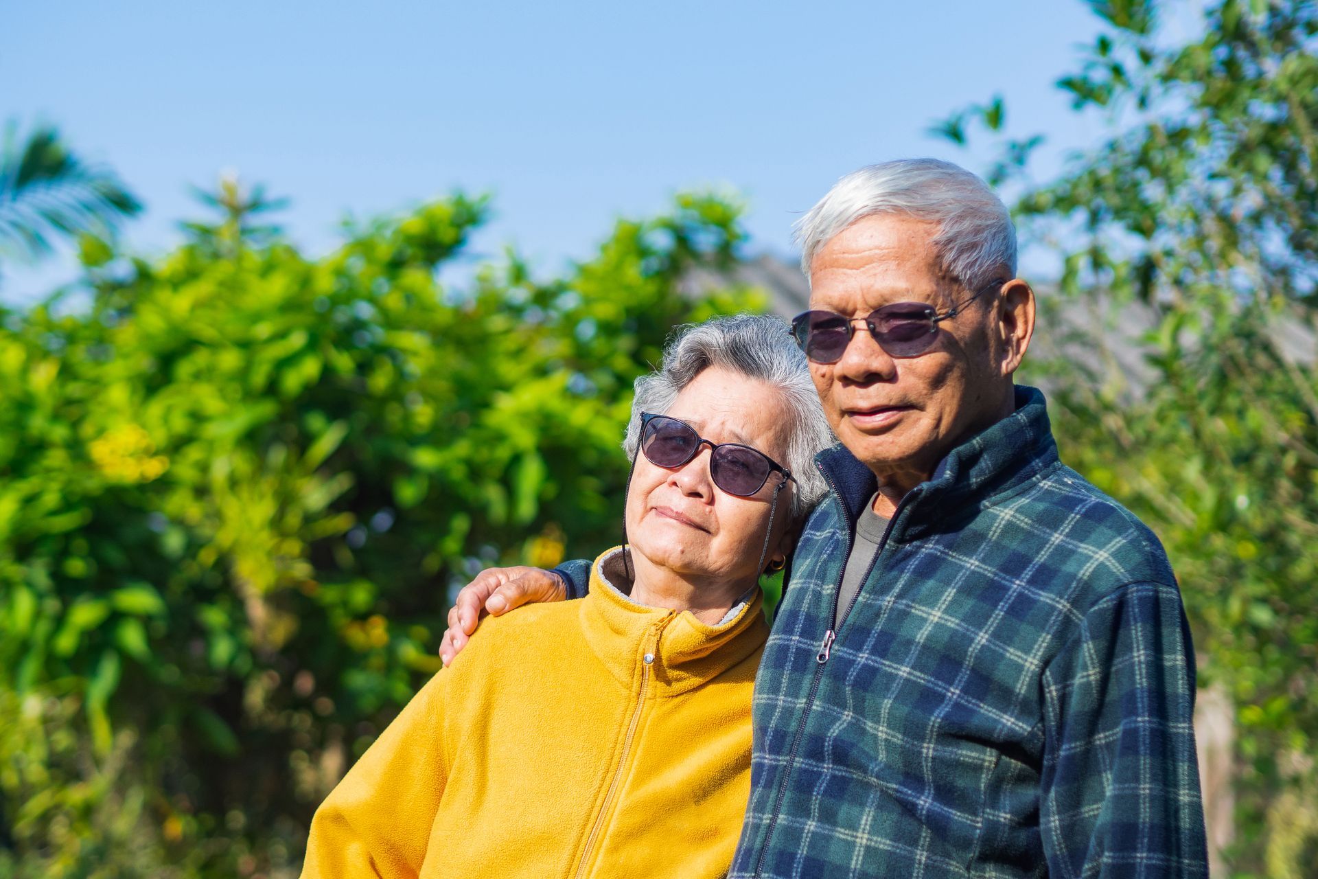 Two people smiling outdoors in sunlight, with green trees in the background.
