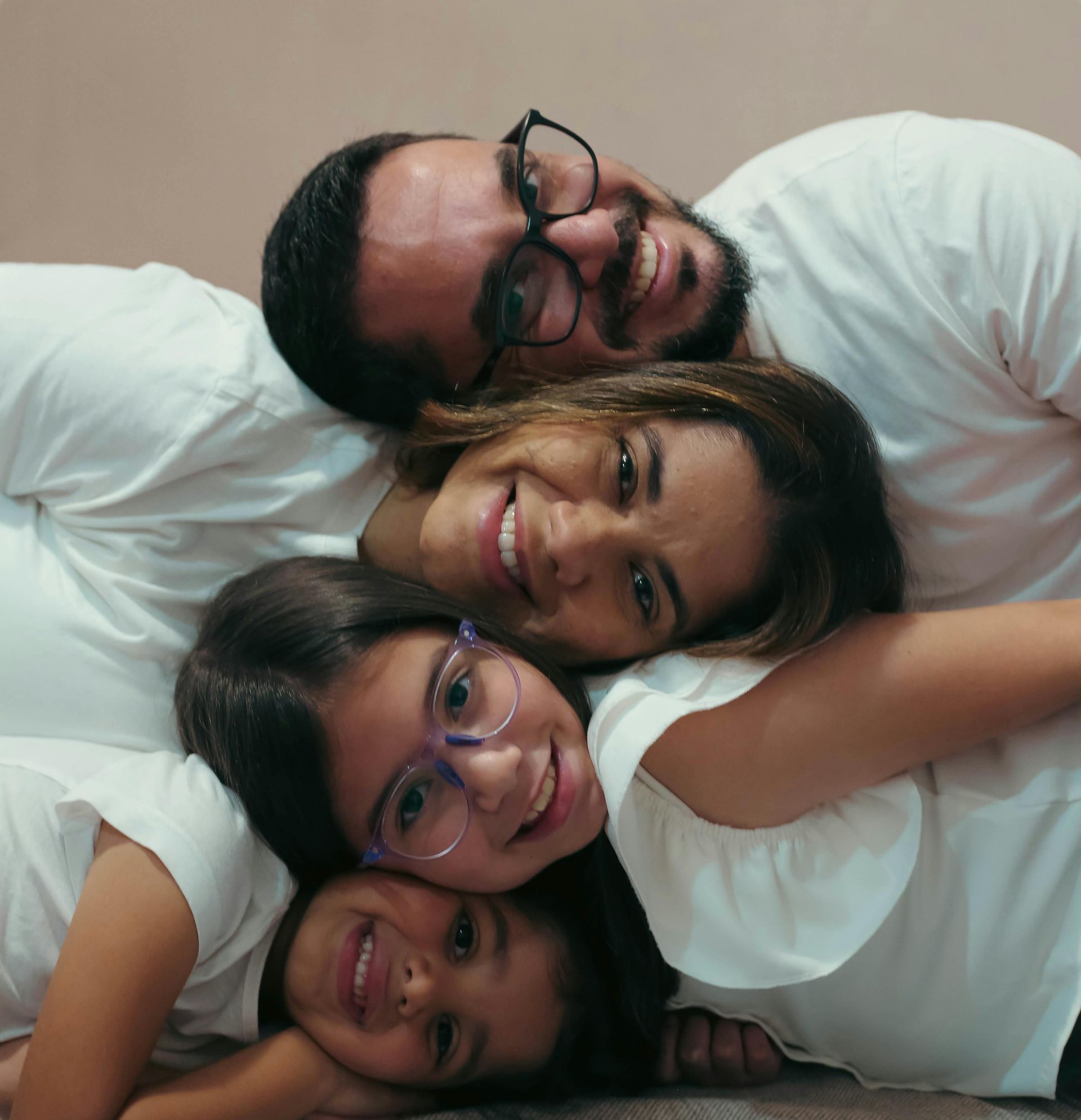 Four smiling people in white shirts lying close together, heads stacked on a bed.