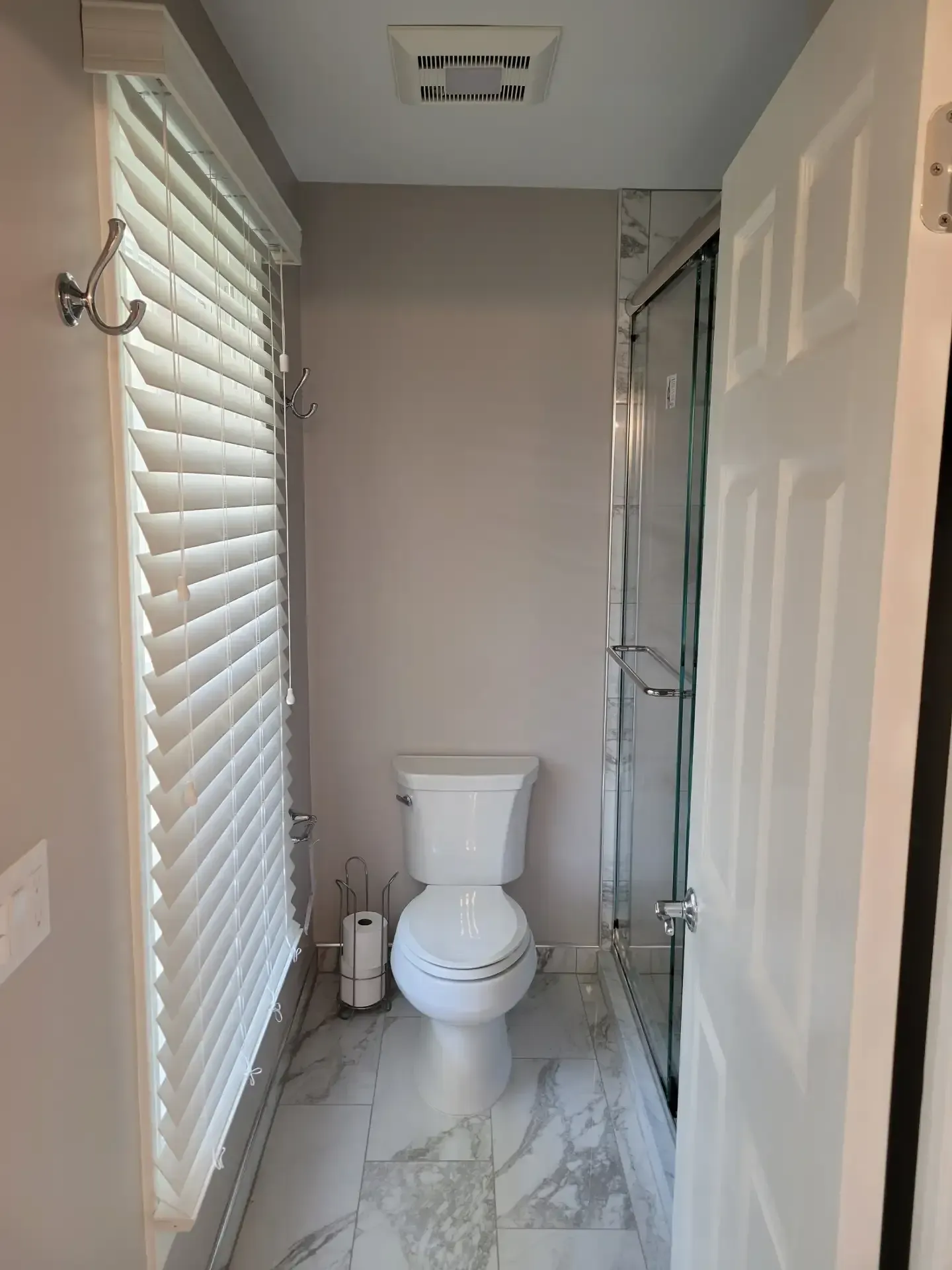 A small, modern bathroom with a toilet, shower, and window with white blinds. Light gray walls and white trim.