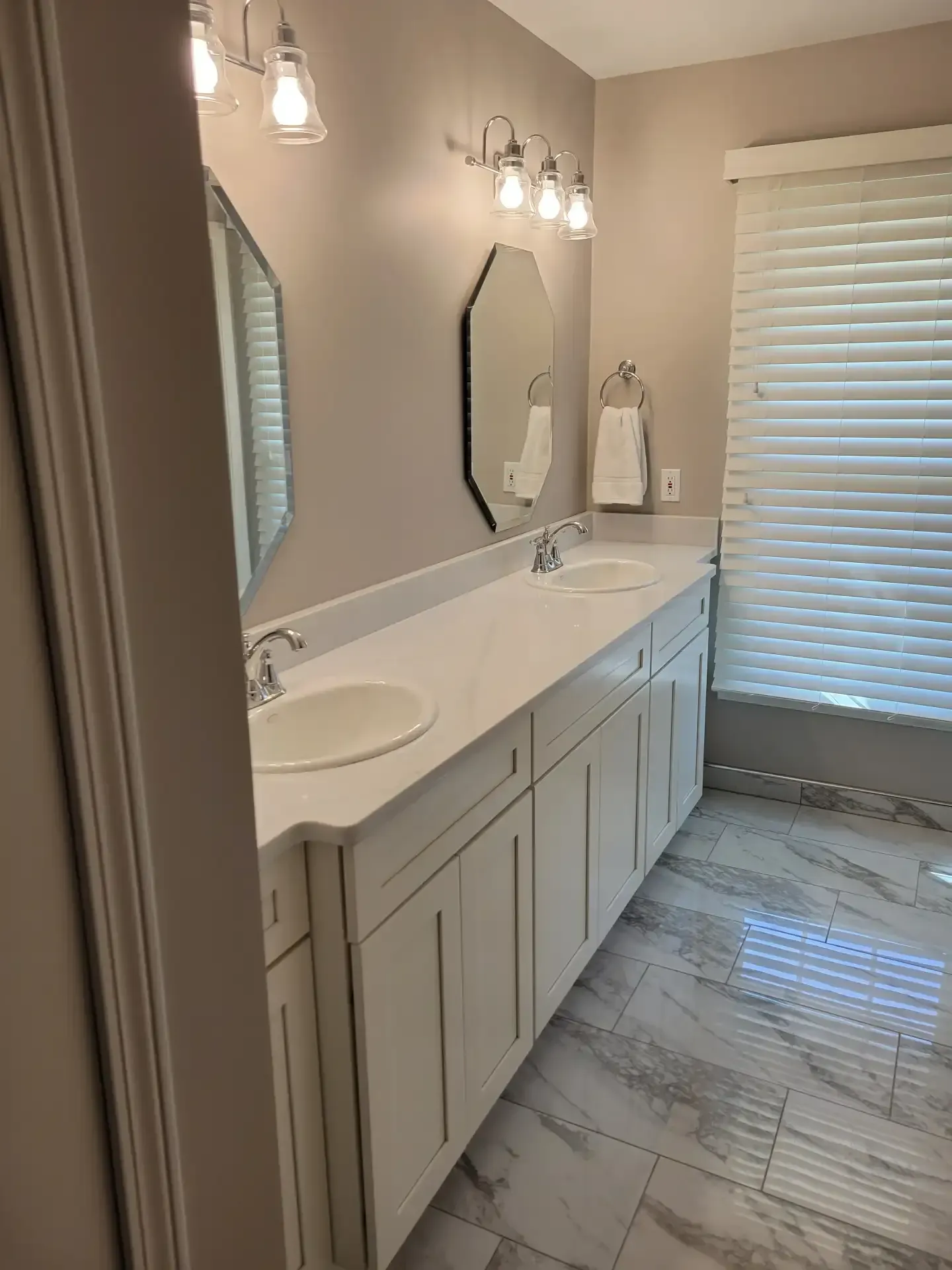 Bathroom with white vanity, two sinks, and octagonal mirror. Gray walls and window with blinds.