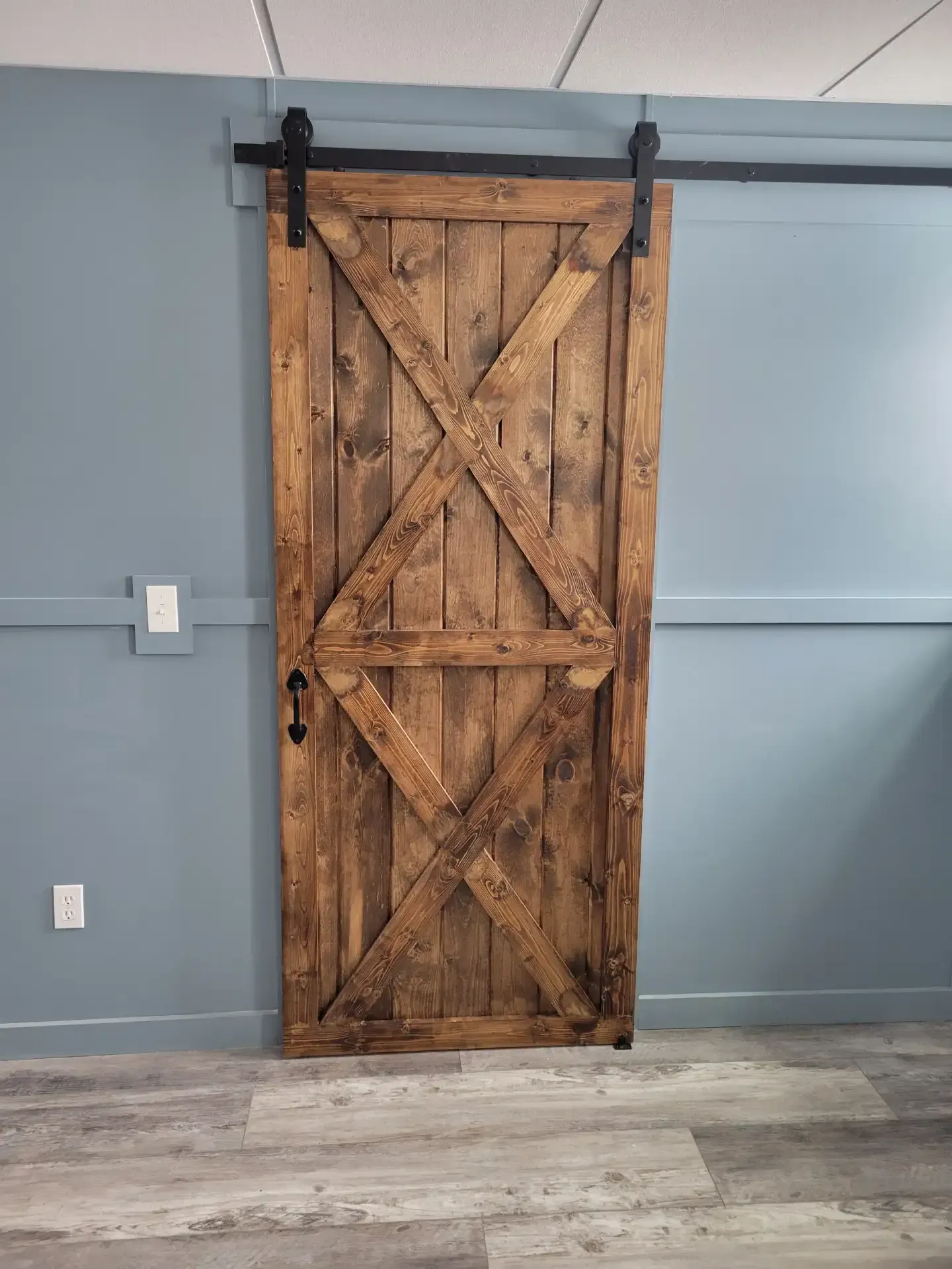 Barn door with X-shaped crossbeams, mounted on a black sliding track, against a blue-gray wall.