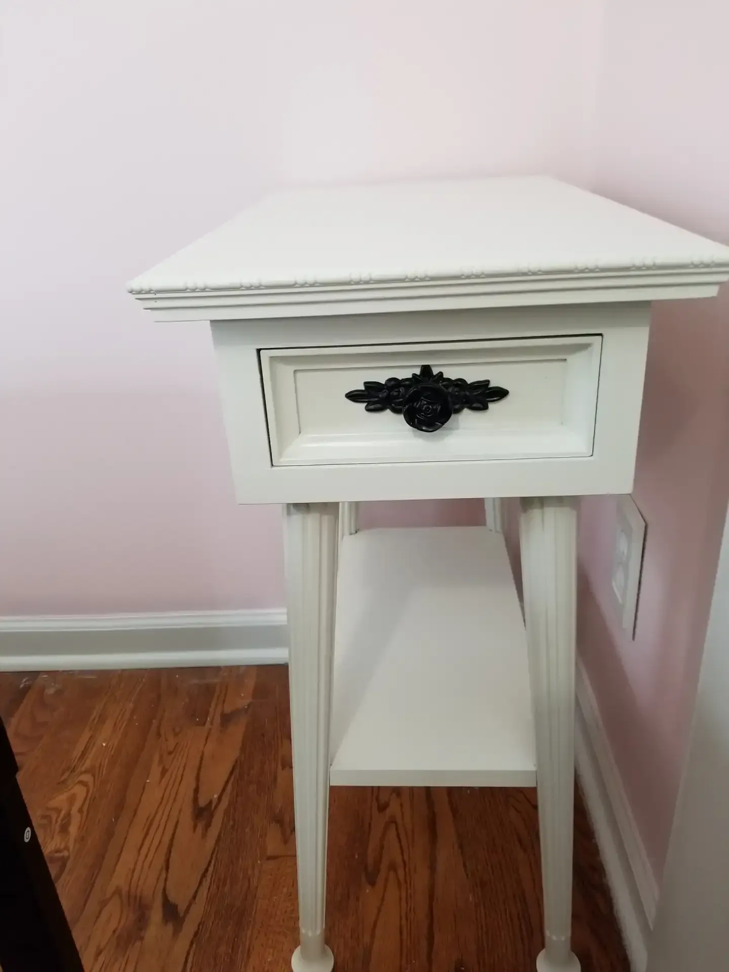 White nightstand with a drawer, shelf, and black handle on a wood floor next to a pink wall.