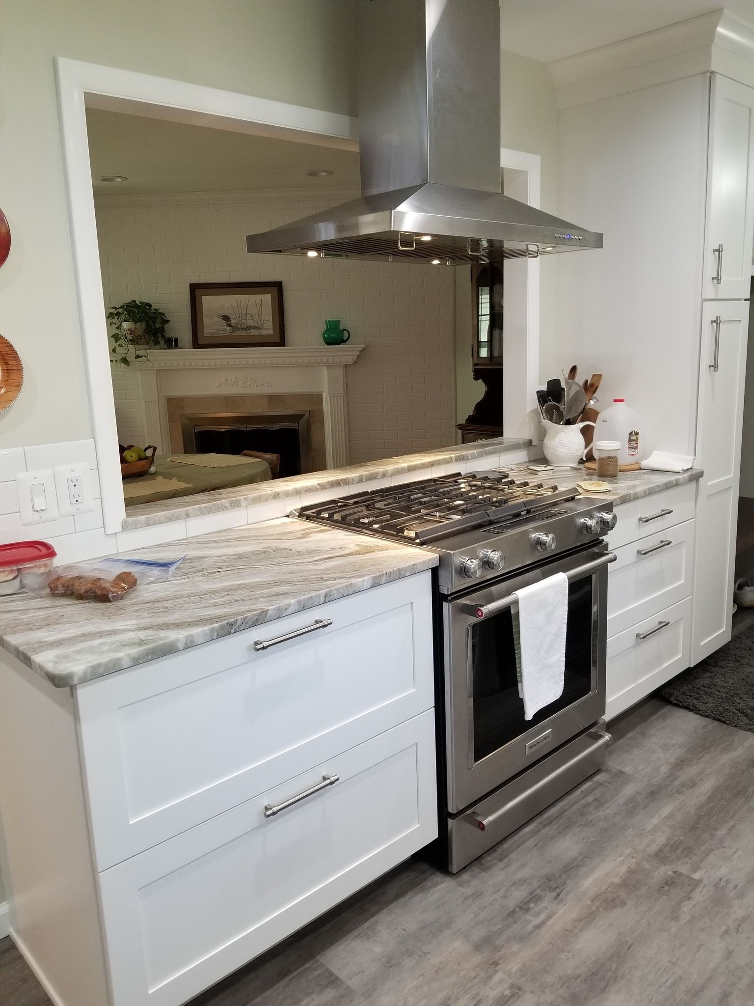 A modern white kitchen with granite countertops, a stainless steel gas range, and a chimney hood overlooking a fireplace.