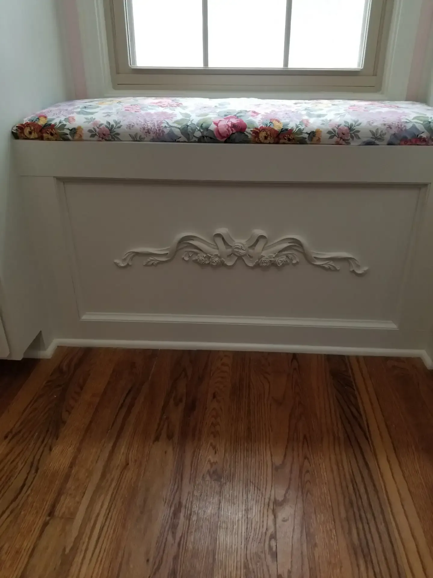 Window seat with floral cushion, decorative molding on white storage unit, set against wooden floor.