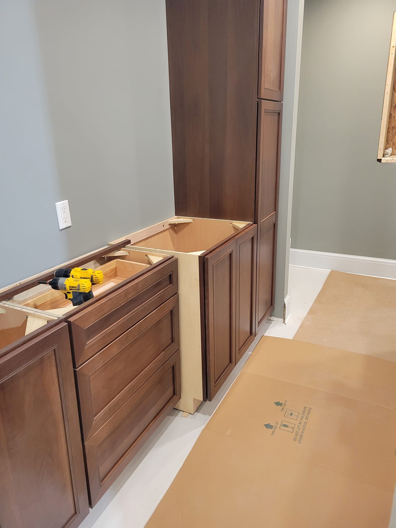 Kitchen cabinets being installed; brown wood, gray wall, construction materials on floor.