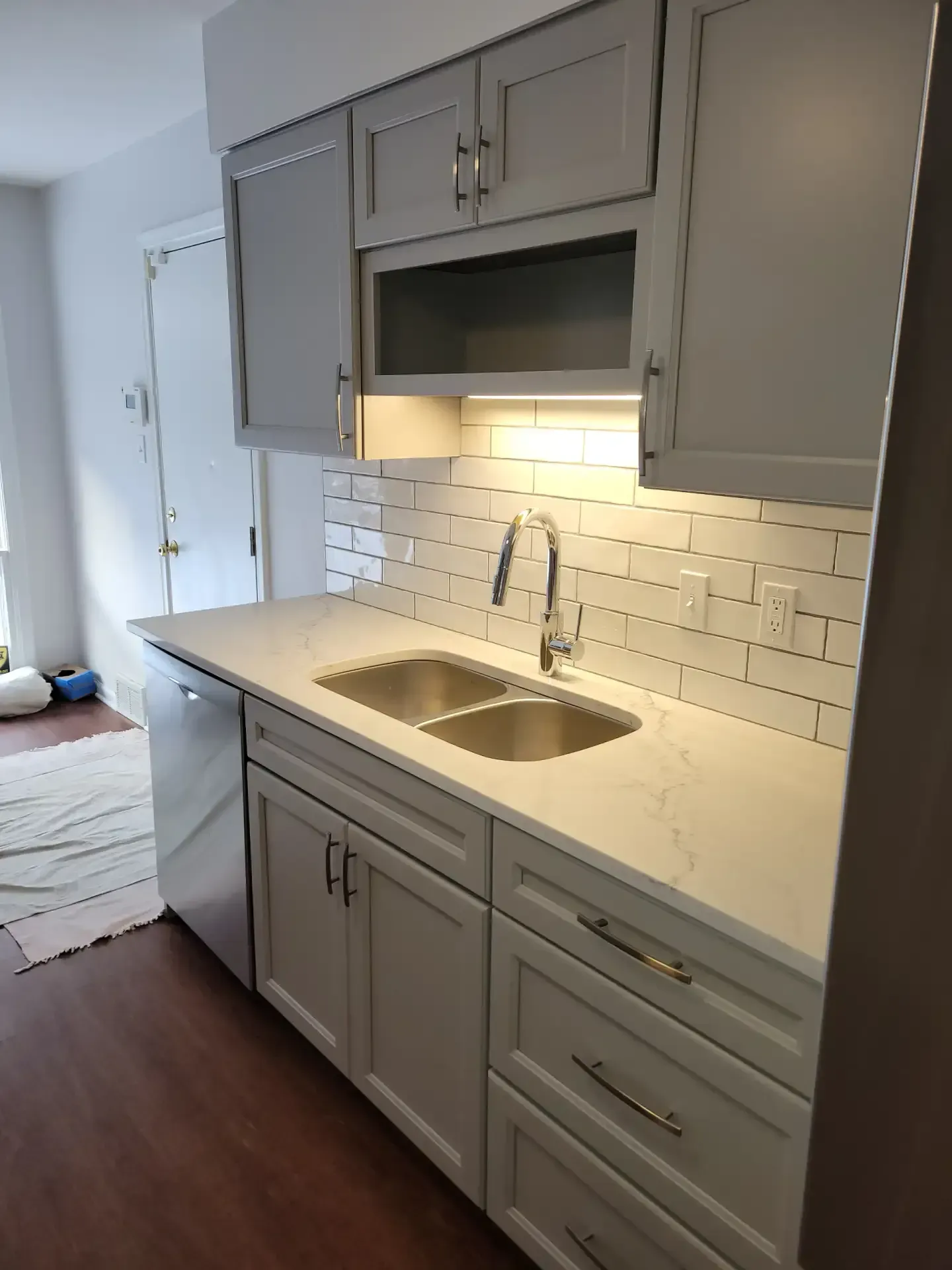 Gray kitchen cabinets with white countertop and subway tile backsplash. Stainless steel sink and faucet.