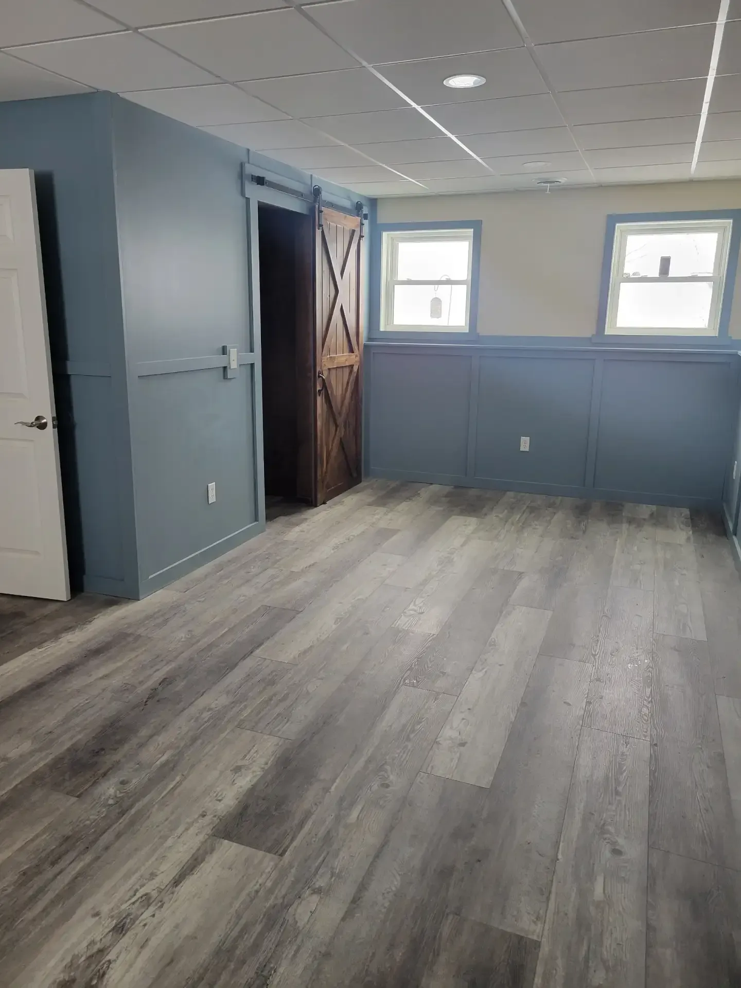 A finished basement with gray wood-look flooring, blue walls, a wooden sliding door, and two windows.