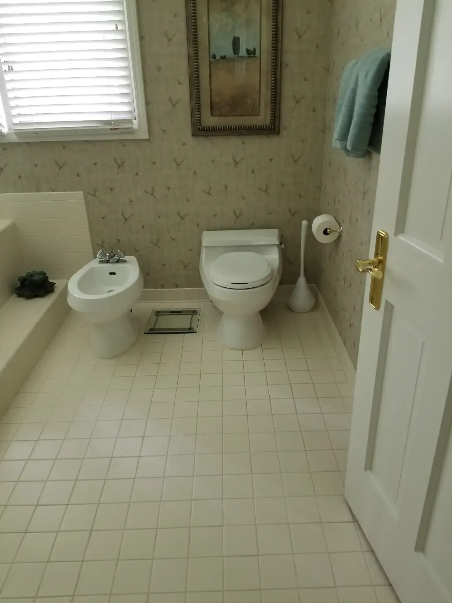 Bathroom with toilet, bidet, and patterned wallpaper. White tiled floor and partially visible door.