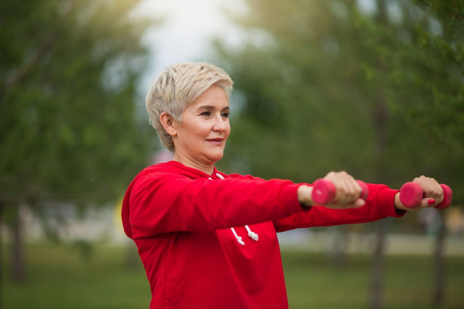 A woman wearing a red hoodie exercises outdoors, holding small pink dumbbells with arms extended forward.