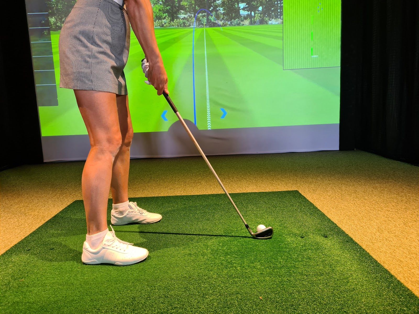 Person about to swing a golf club inside, facing a screen showing a golf course.