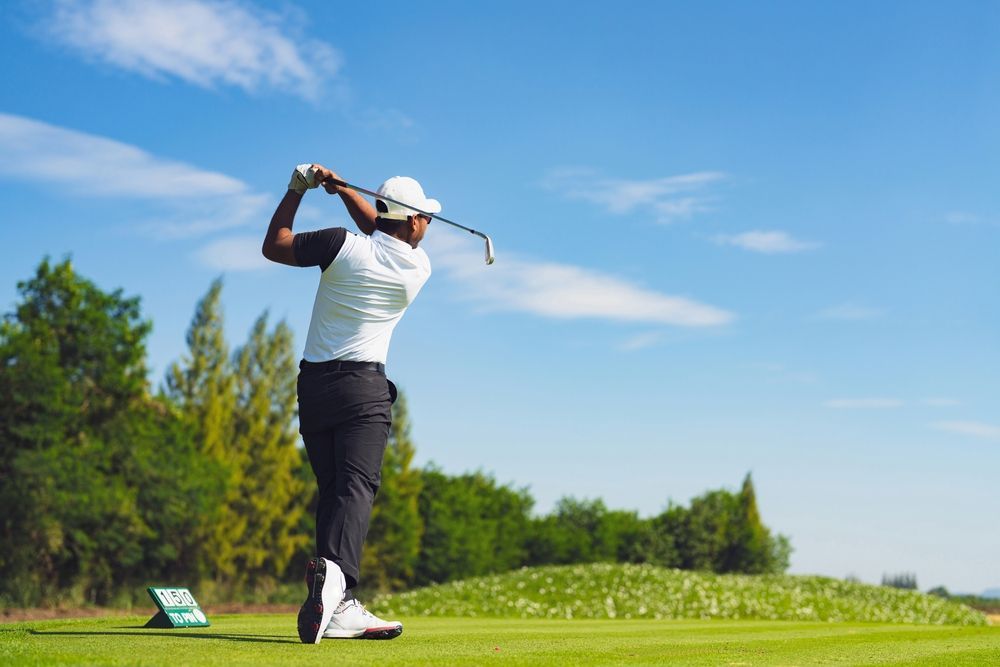 A golfer follows through on a swing on a sunny green course, with trees in the background and a clear blue sky.