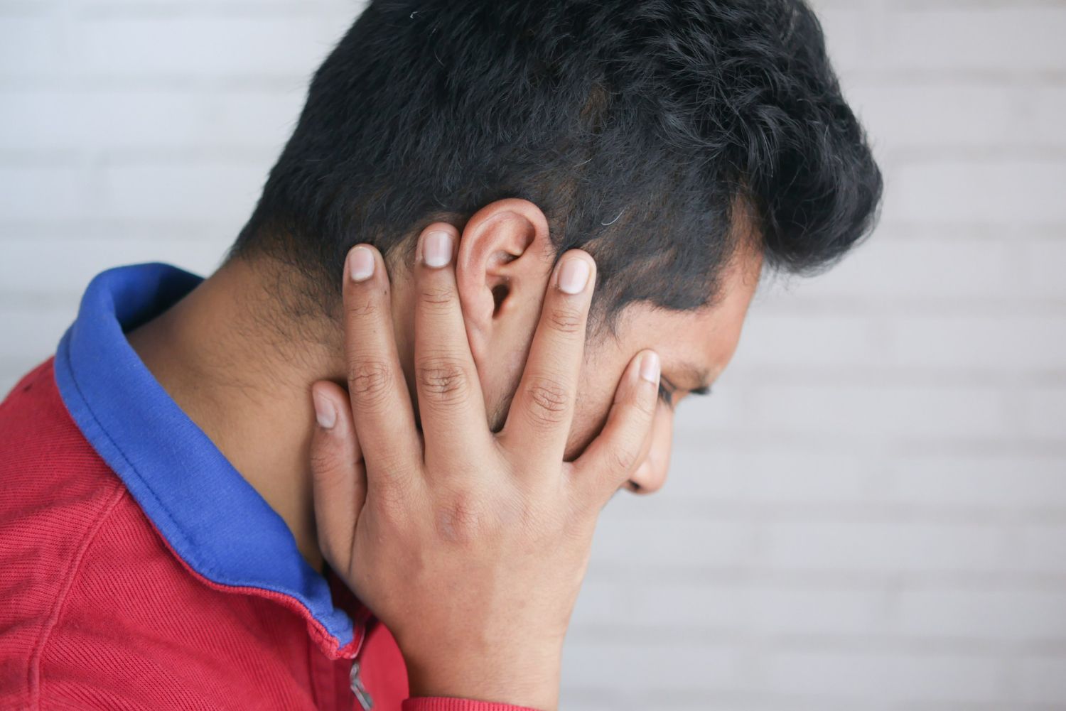A person wearing a red shirt with a blue collar, holding their hand against the side of their head as if in pain.