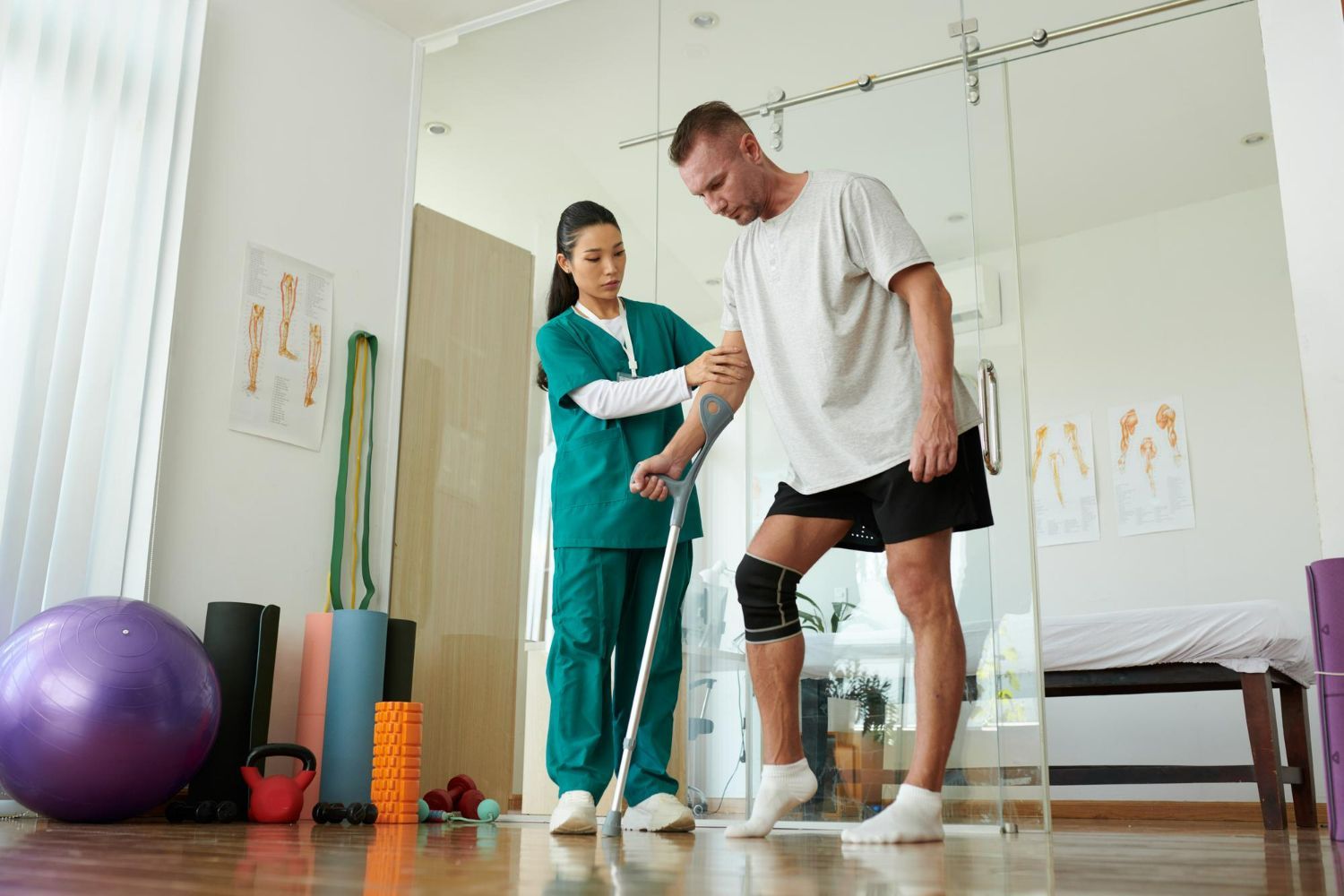 A nurse is helping a man walk with crutches in a gym.