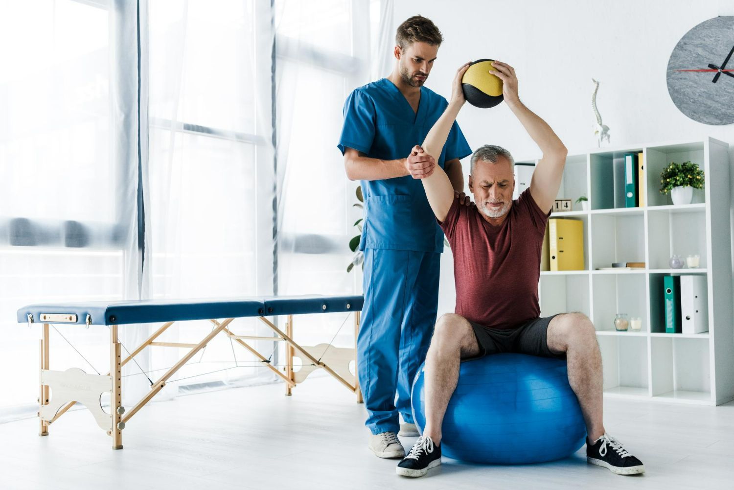 Physical therapist assisting a patient on a blue exercise ball in a bright clinic room