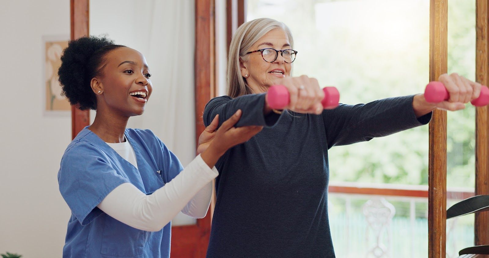 Woman exercising with weights, guided by a healthcare professional indoors. Both are smiling.