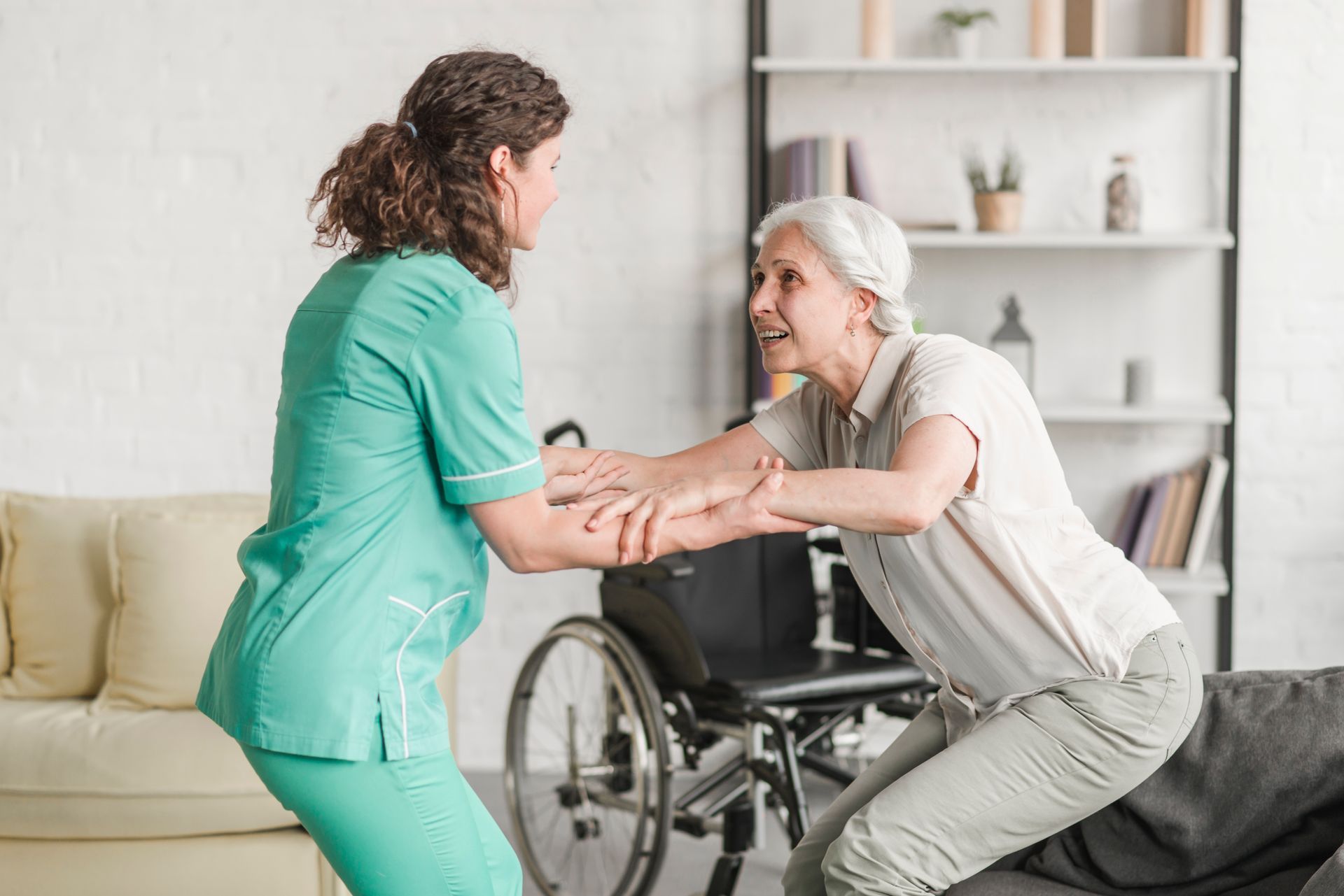 Nurse assisting an elderly woman from wheelchair. They're in a living room, woman's arm held.