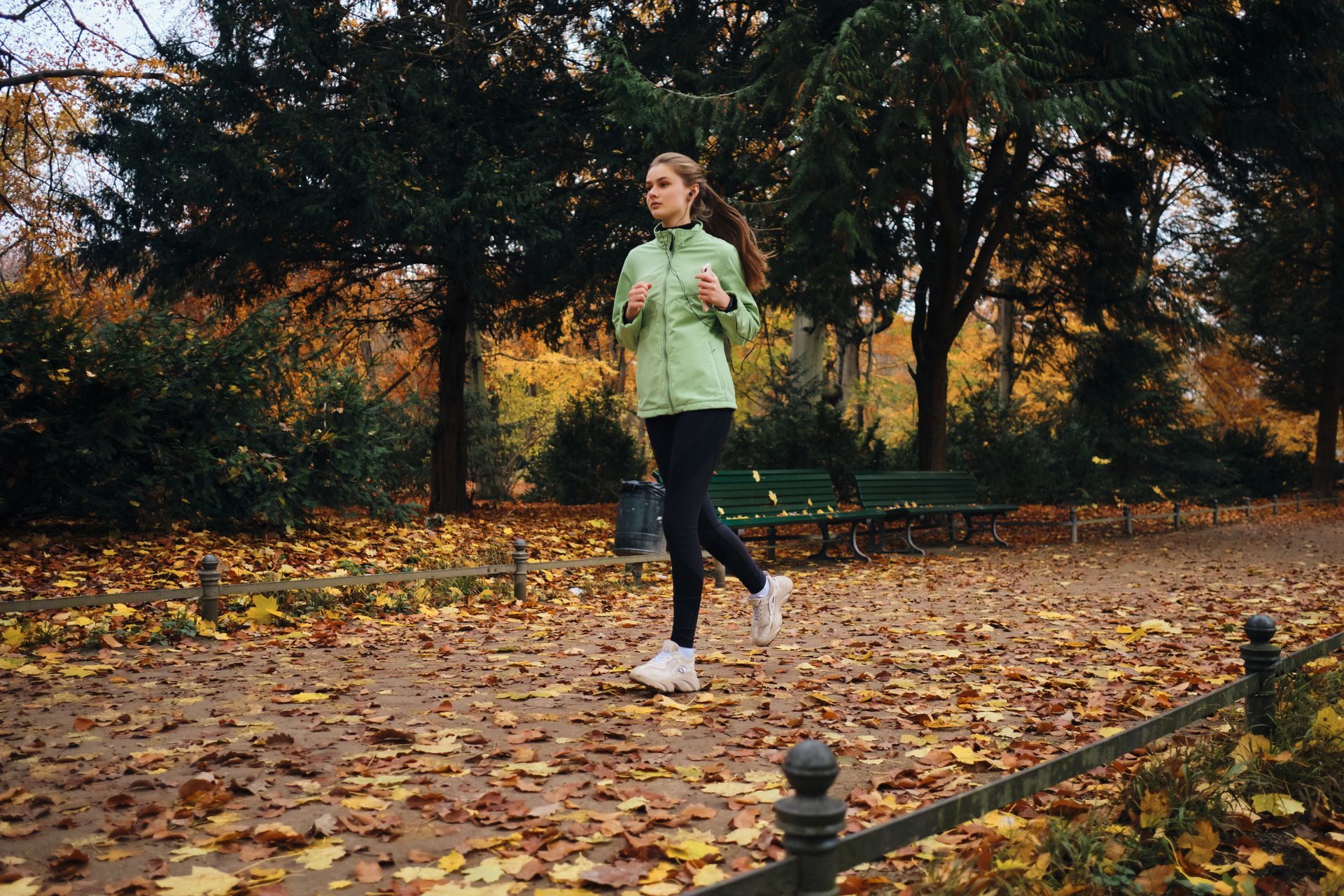 Woman jogging in a park with autumn leaves, wearing a green jacket and black leggings.