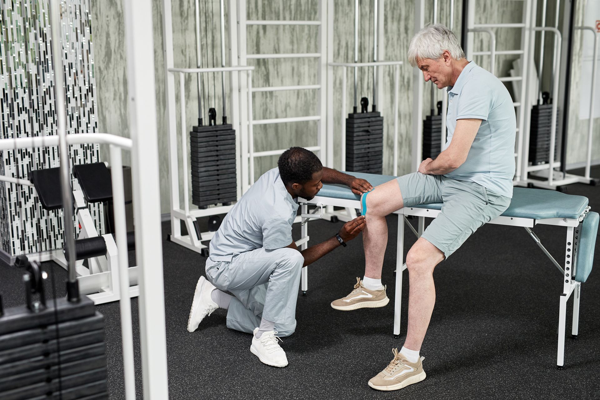 Physical therapist examines senior man's knee in a rehab gym. Black PT kneels, patient sits, both light-skinned.