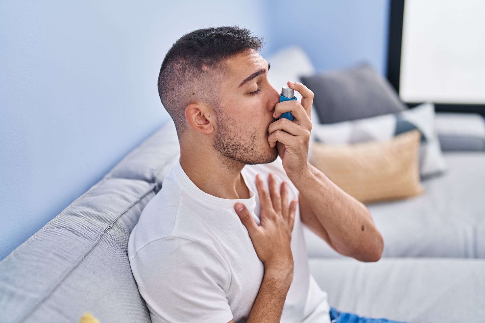 Man using an inhaler, sitting on a couch, holding chest, eyes closed.