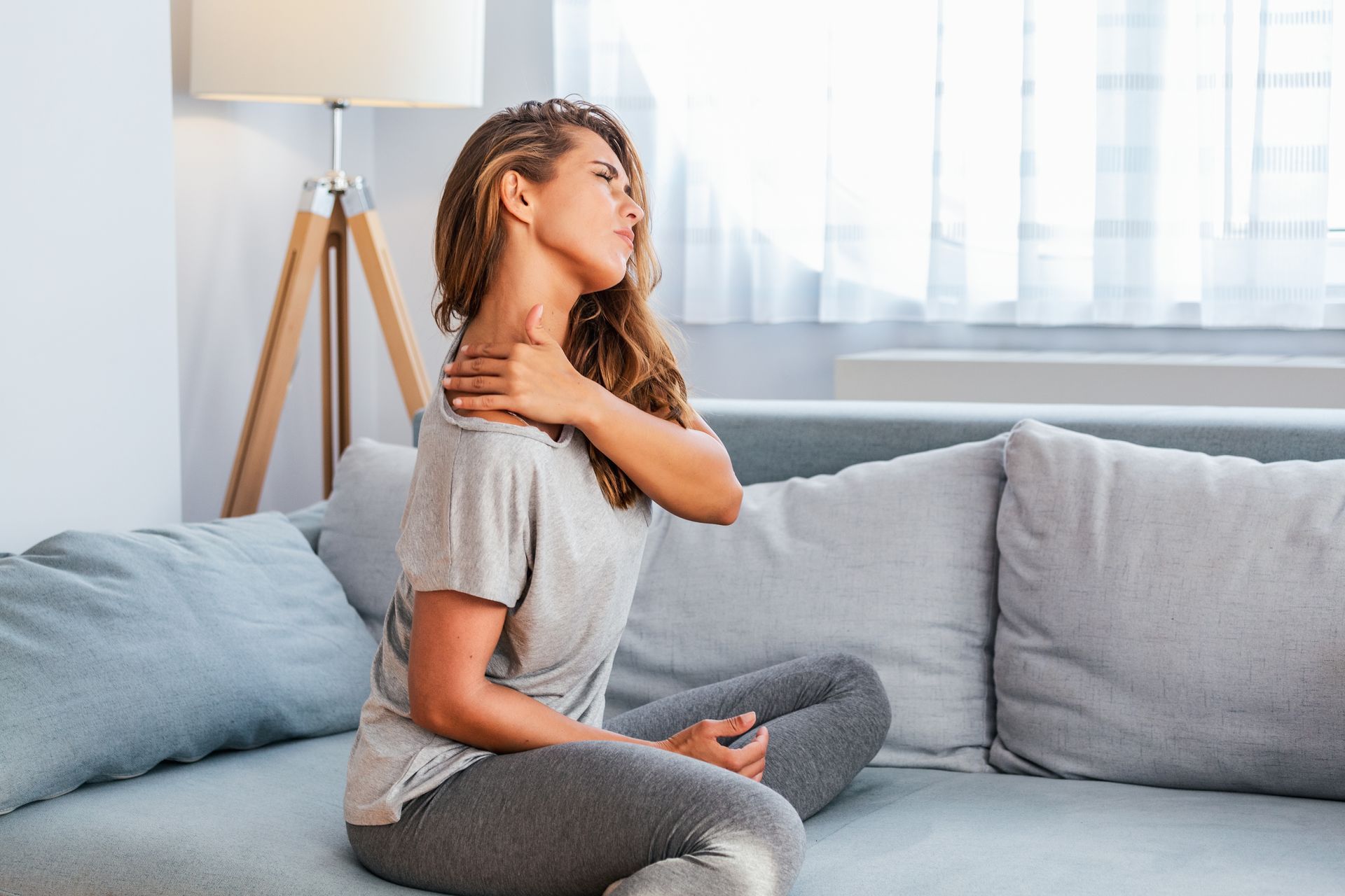 Woman seated on couch clutches shoulder, showing pain. Indoors with lamp and window.