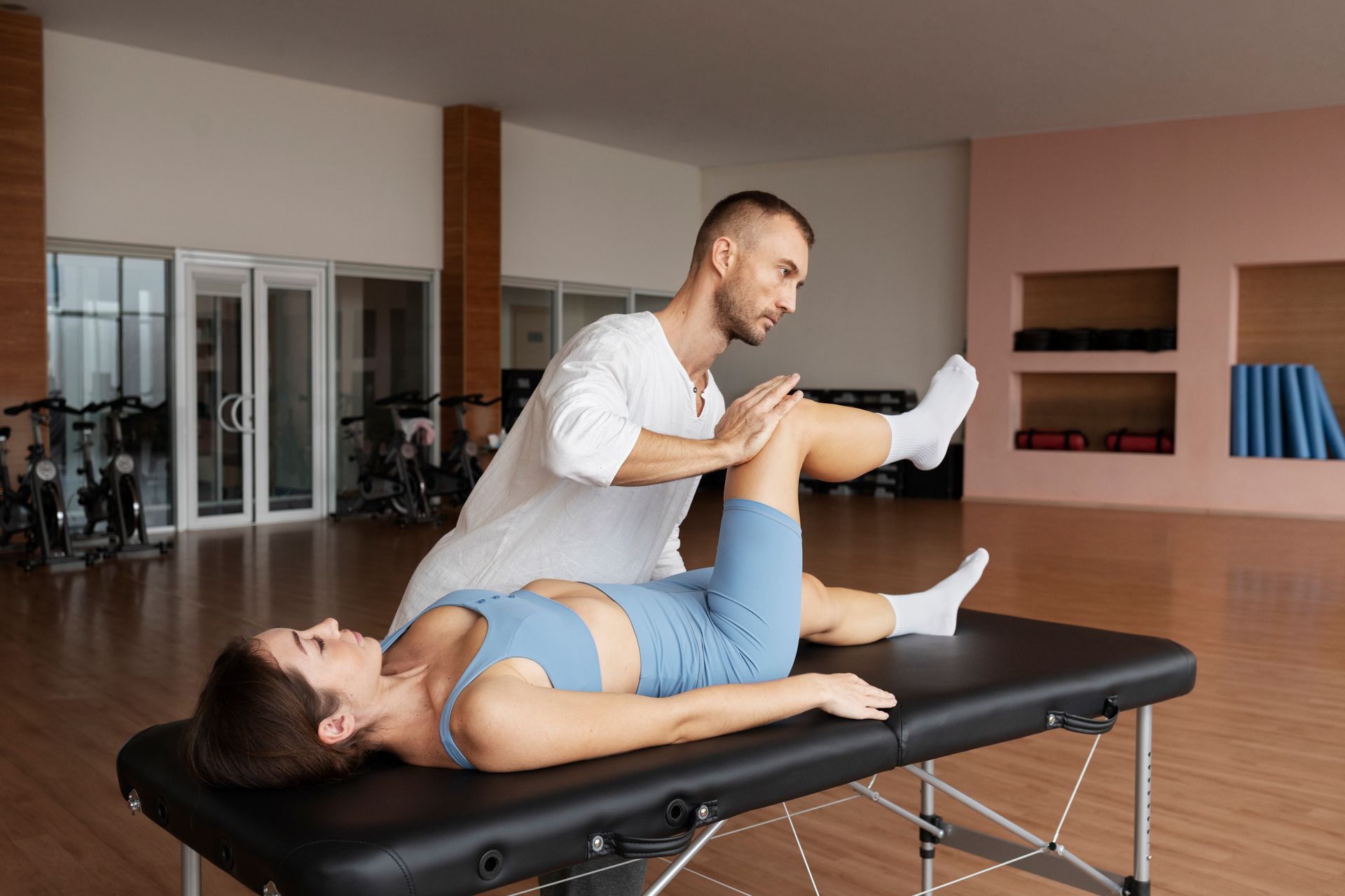 A man is stretching a woman 's leg on a table in a gym.