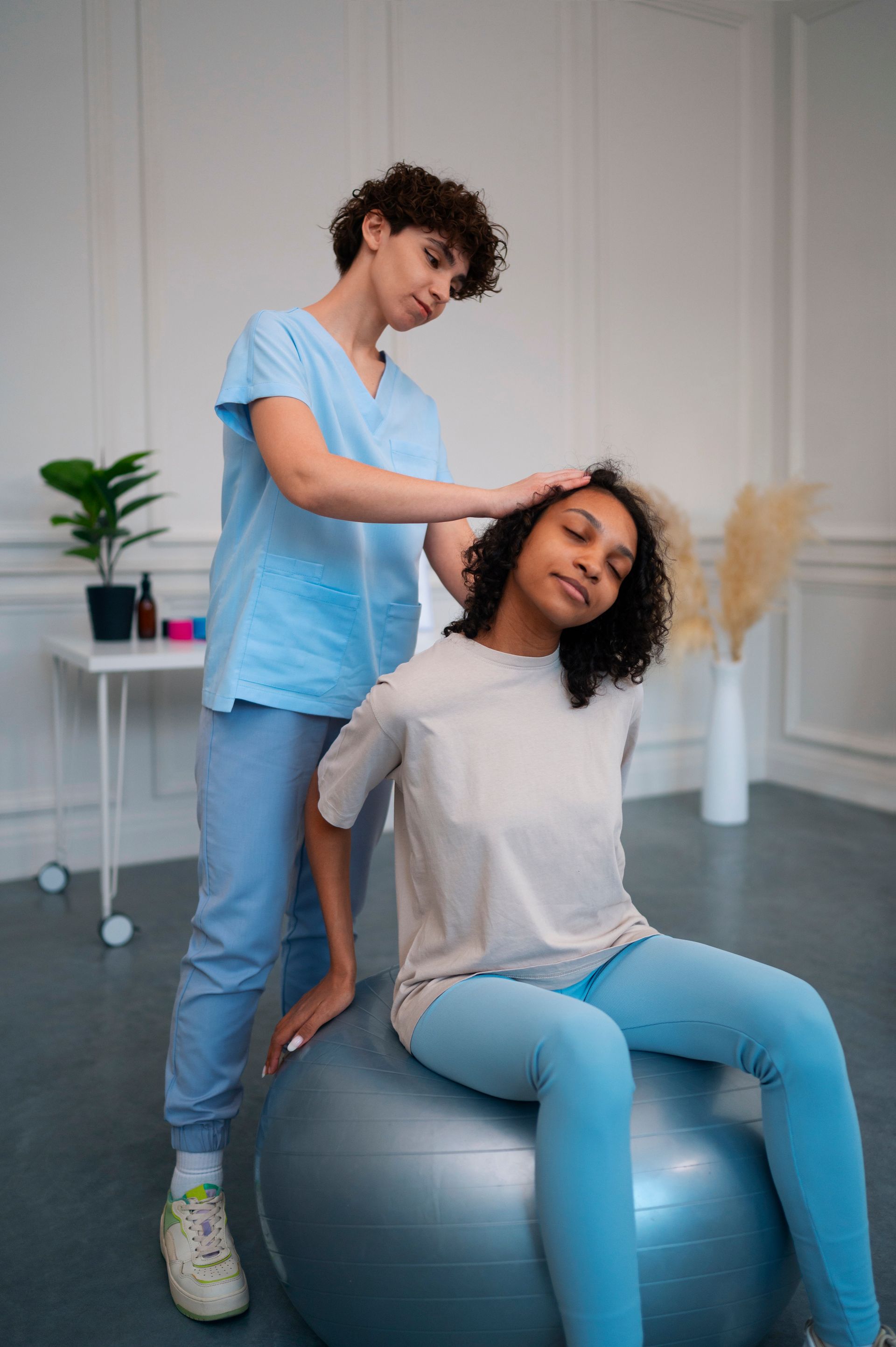 A woman is sitting on an exercise ball while a woman is standing behind her.