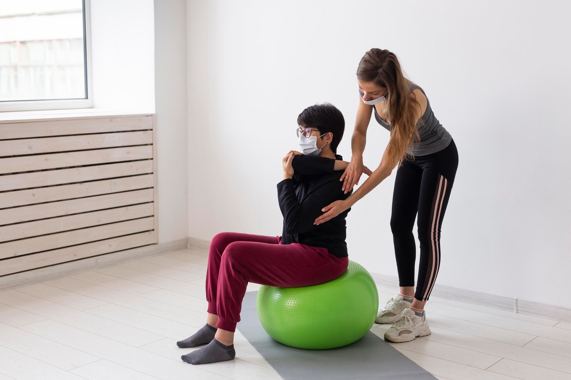 A woman wearing a mask is helping a woman sit on a green exercise ball.