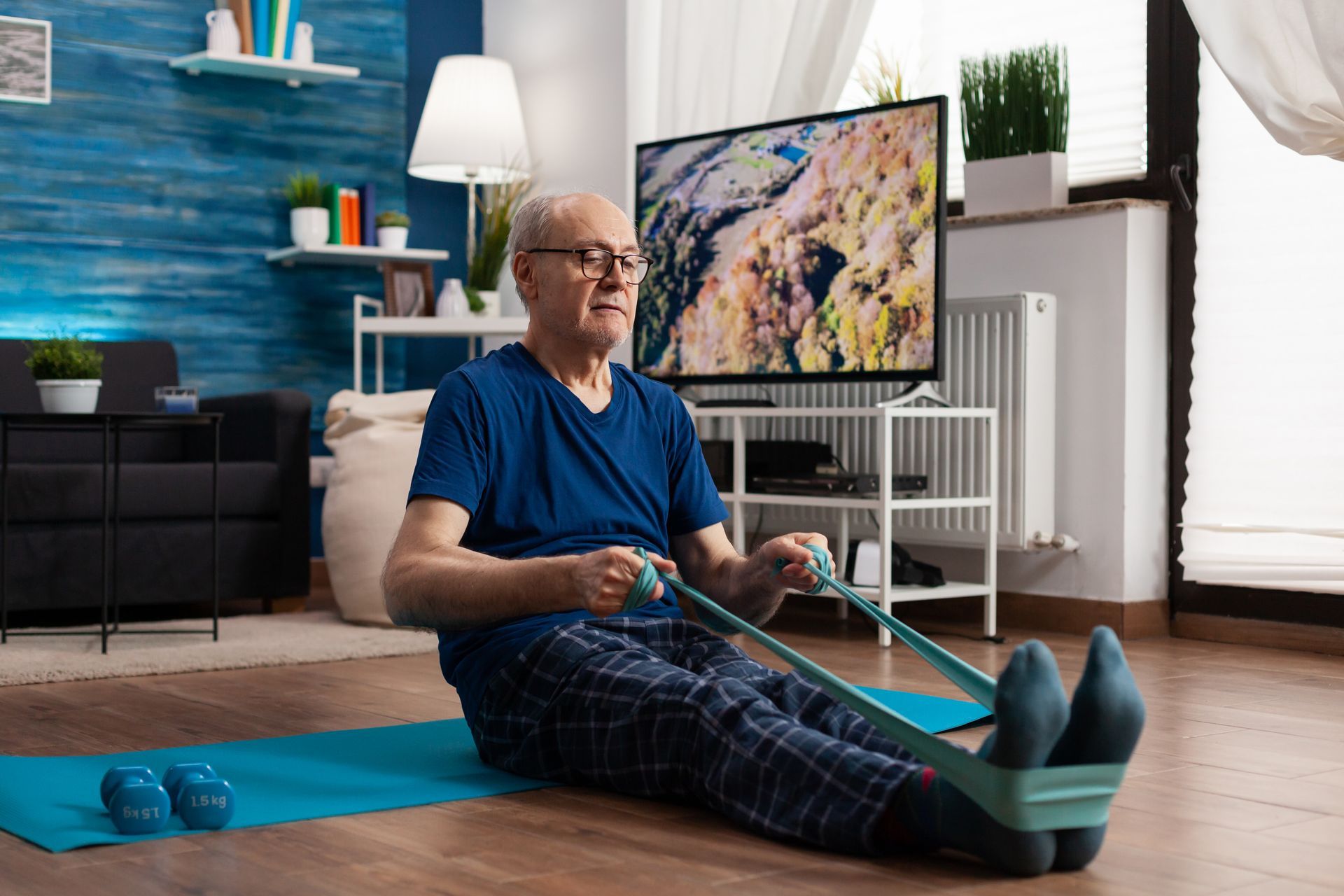 An elderly man is sitting on a yoga mat doing exercises with a resistance band.