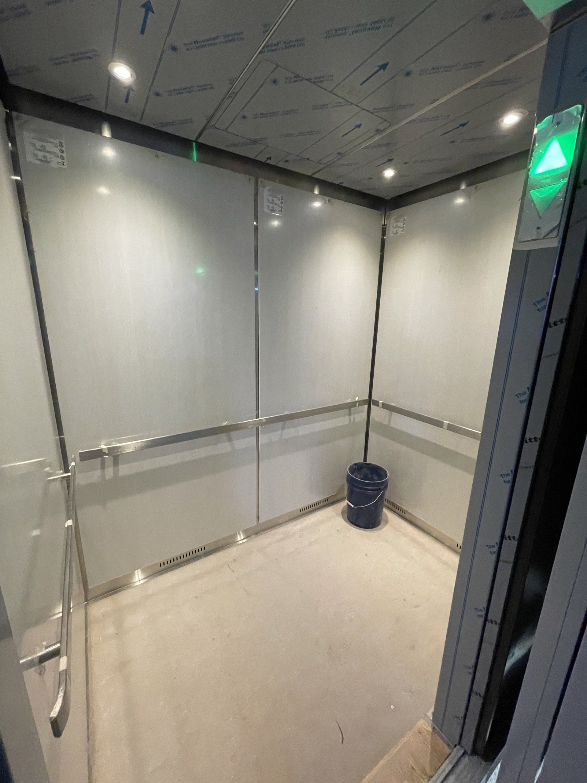 Intérieur d'ascenseur vide avec des murs gris, un sol et une flèche verte vers le haut.