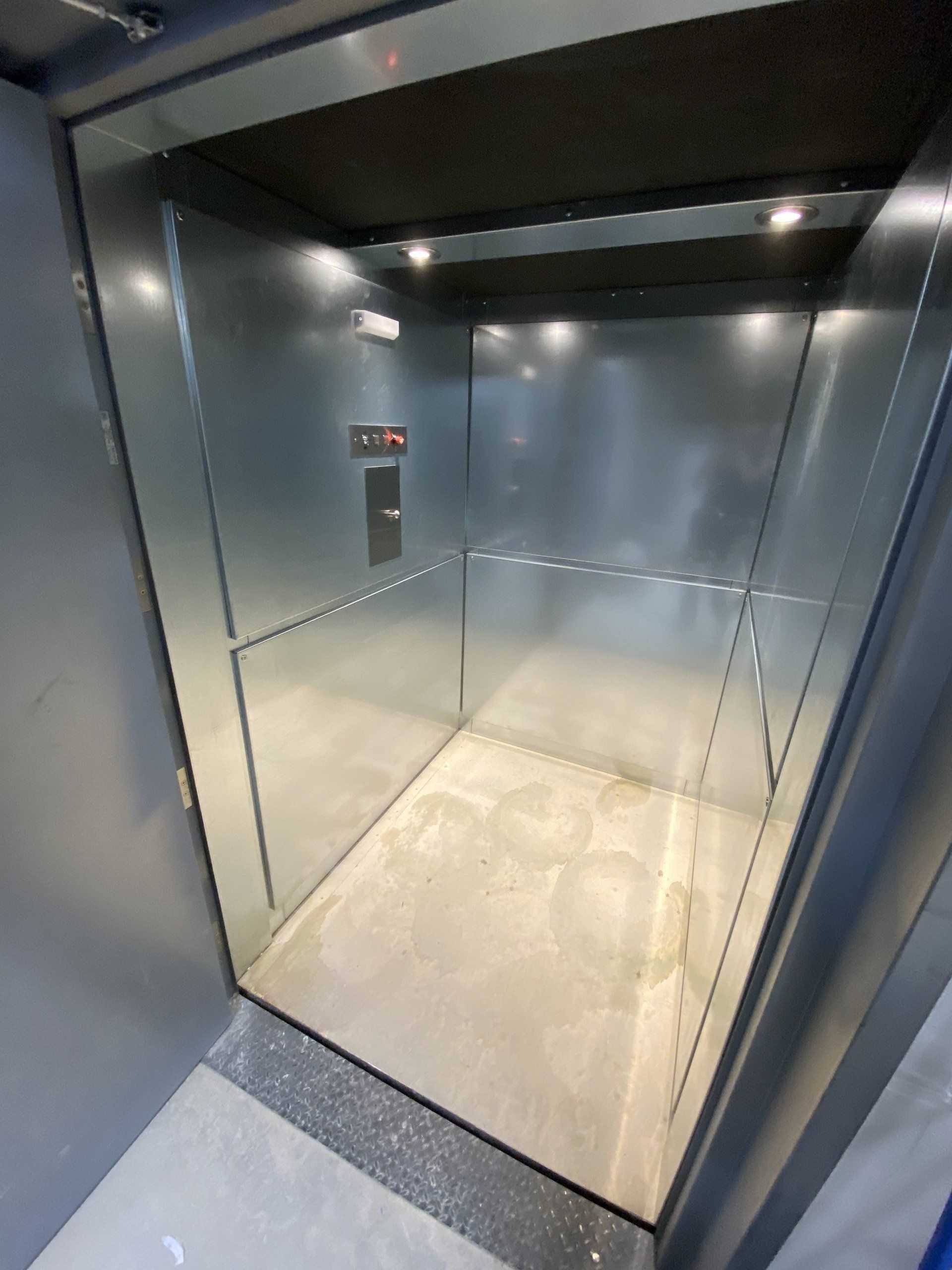 Intérieur d'un ascenseur aux murs argentés, au plafond et au sol sombres. Panneau de commande visible.