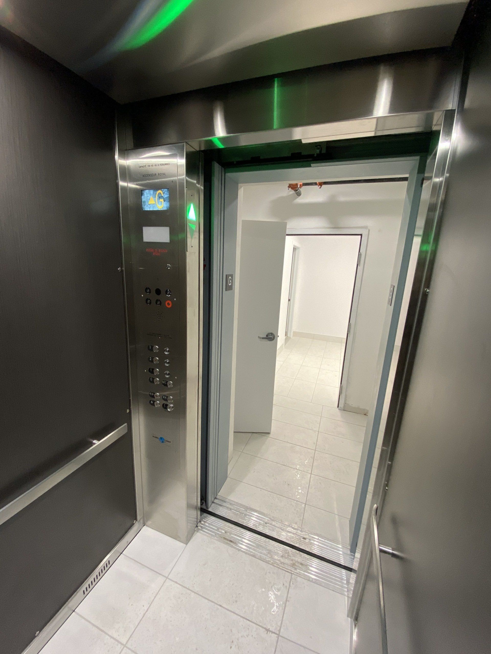 Intérieur d'un ascenseur, portes ouvertes, révélant un couloir blanc. Panneau en acier inoxydable avec boutons.