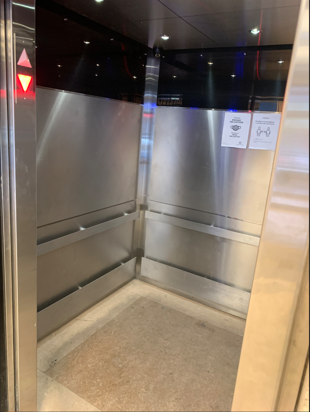 Intérieur d'ascenseur vide avec parois en acier inoxydable, rampes et moquette. Flèche rouge vers le haut allumée.