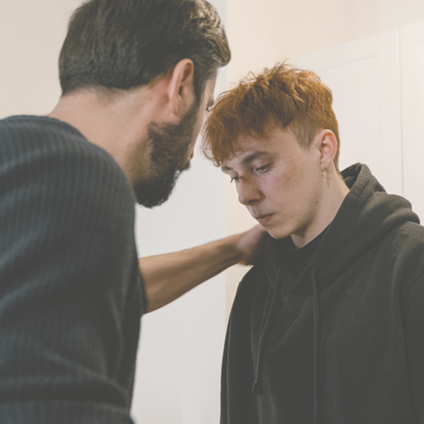 Man comforting a young person, both indoors. The man's hand rests on the younger person's shoulder.