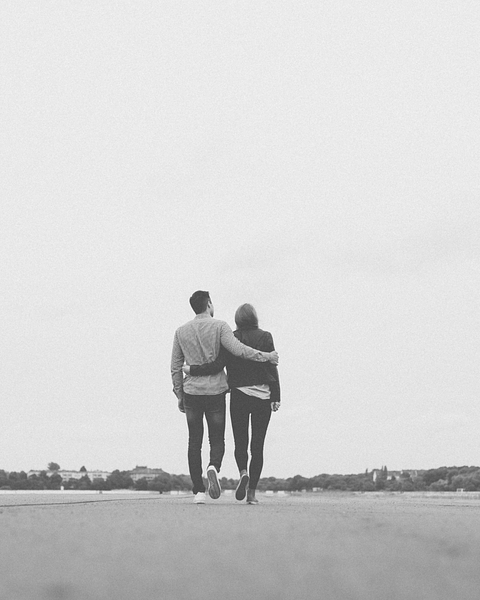 Couple walking away arm in arm across a gray surface, with a distant treeline and sky.