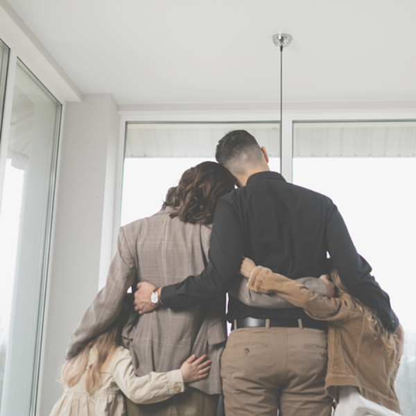 Family of four embraces, looking out a window. Neutral tones, modern interior.