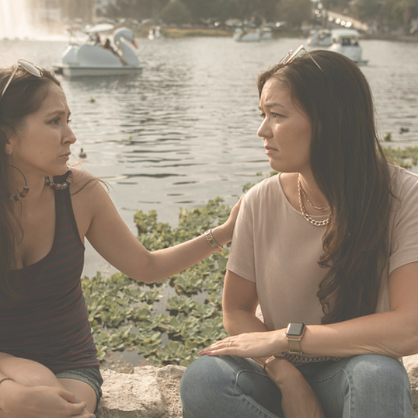 Two women seated by a lake, one consoles the other, watercraft in the background.