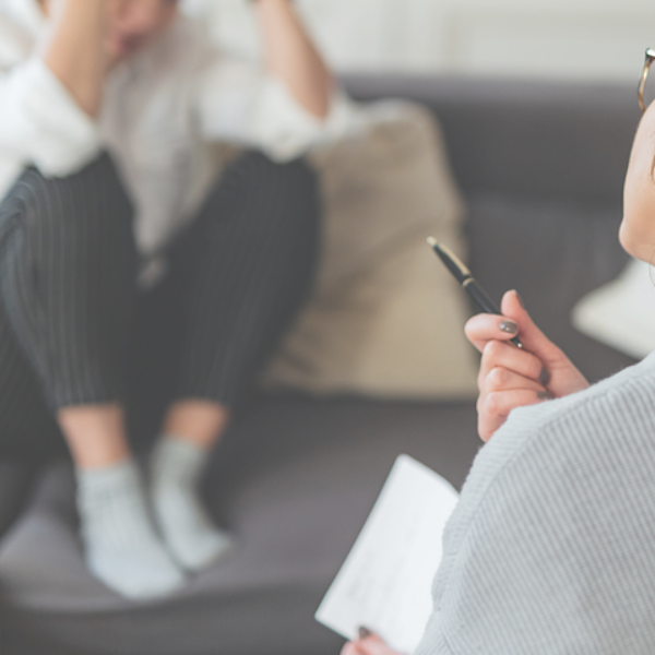 Therapist and patient in a counseling session; patient appears distressed, holding head; therapist holds pen and paper.