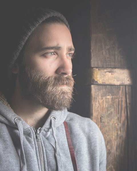 Man with a beard in a gray hoodie and hat, looking off to the side near a wooden door.