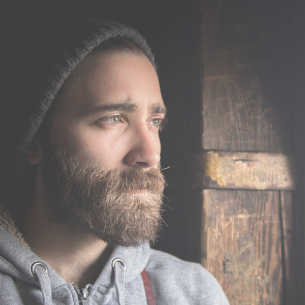 Man with a beard and beanie looking out a window, wearing a gray hoodie.