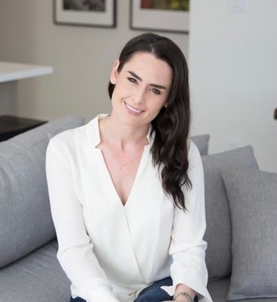 Woman with dark hair smiles, sitting on a grey couch, wearing a white blouse and jeans.