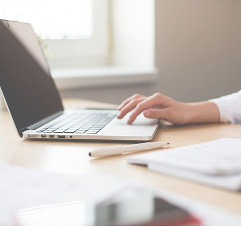 Person's hand on laptop touchpad, working at a desk with a pencil and papers.