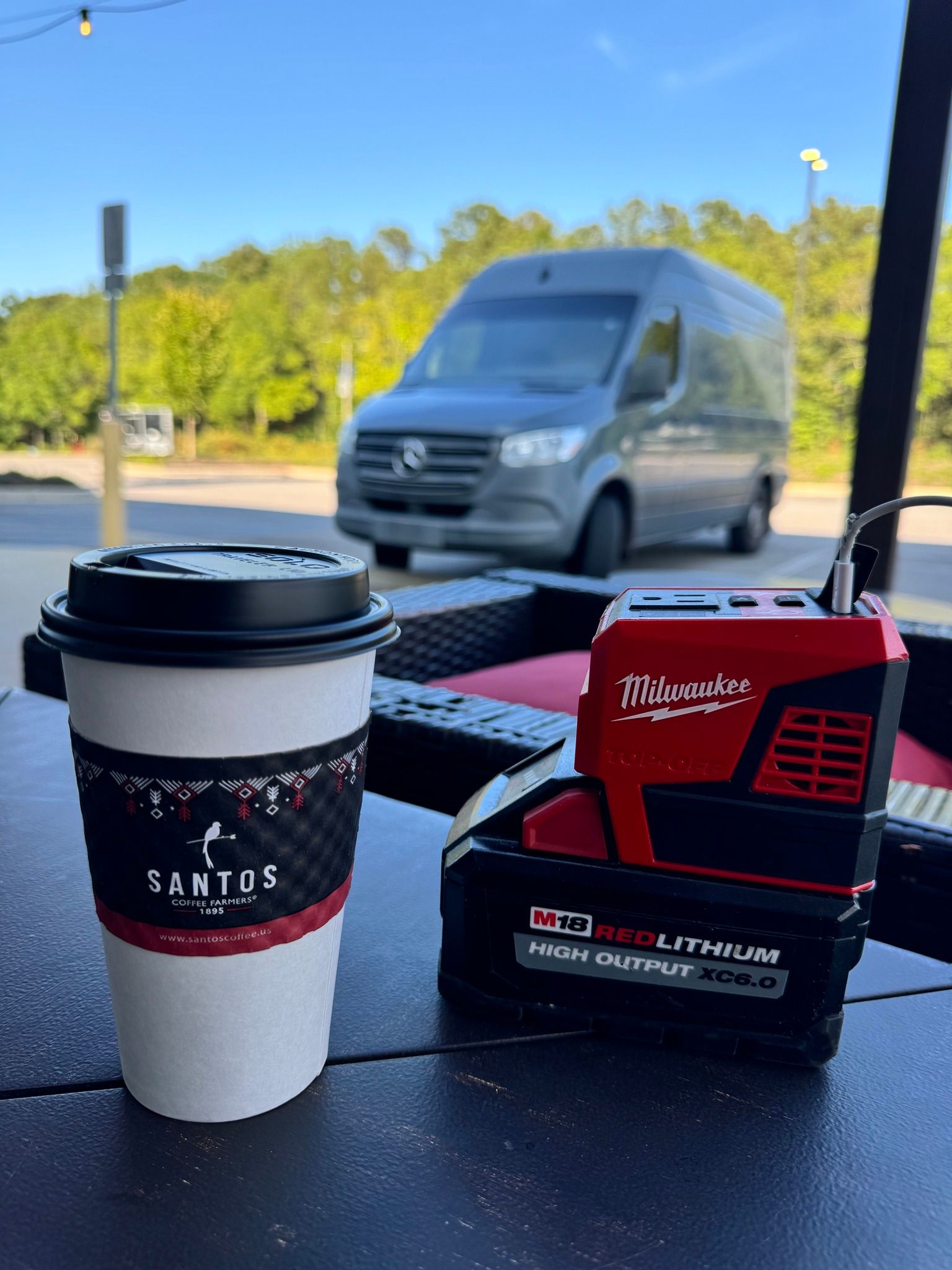 Coffee cup and Milwaukee tool battery on a table with a van in the background.