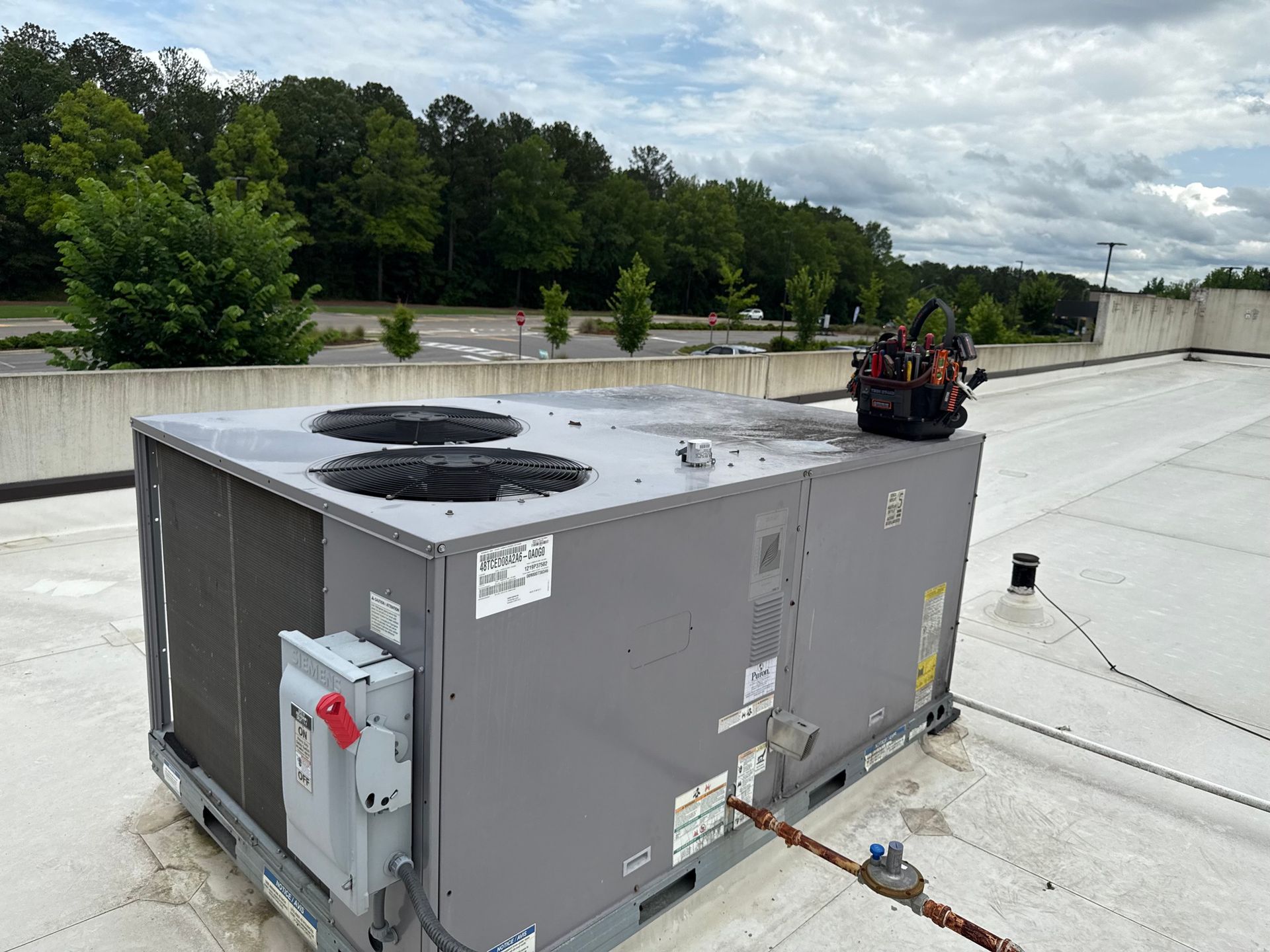 HVAC unit on a rooftop with tools beside it; trees and sky in background.