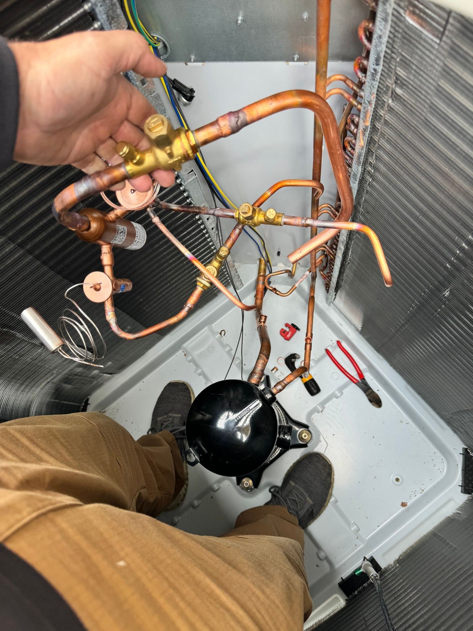 Person working on HVAC components, copper tubing, and a black compressor inside a unit.