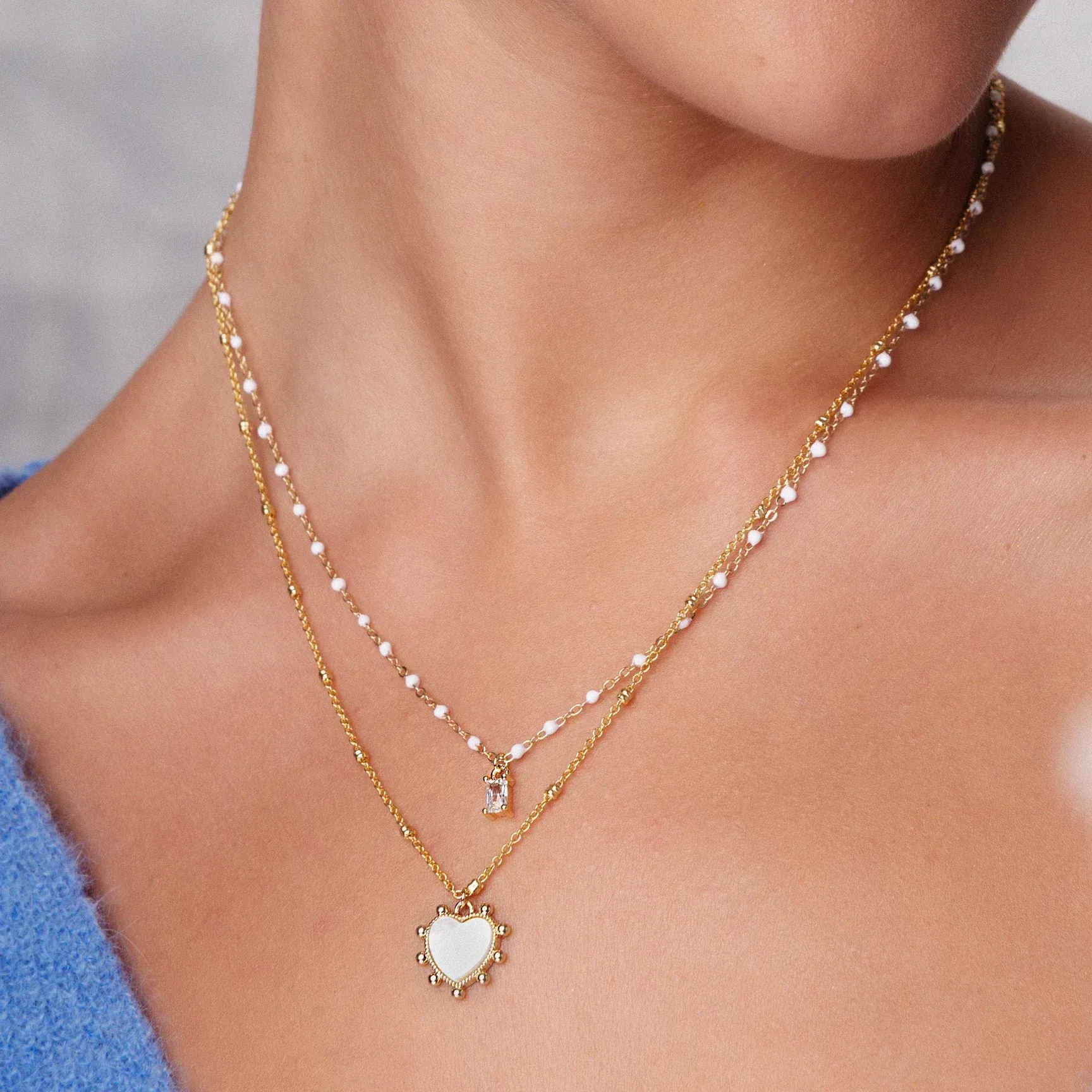 A woman is wearing a gold necklace with pearls and a white heart pendant.