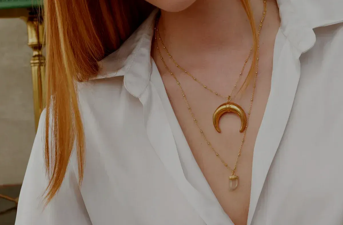 A woman wearing a white shirt and a gold necklace with a crescent moon pendant.