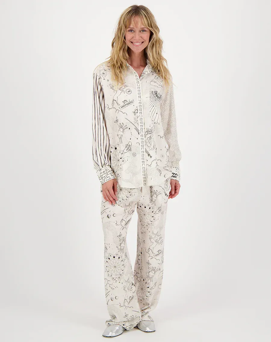 A woman wearing a white shirt and pants is standing in front of a white background.